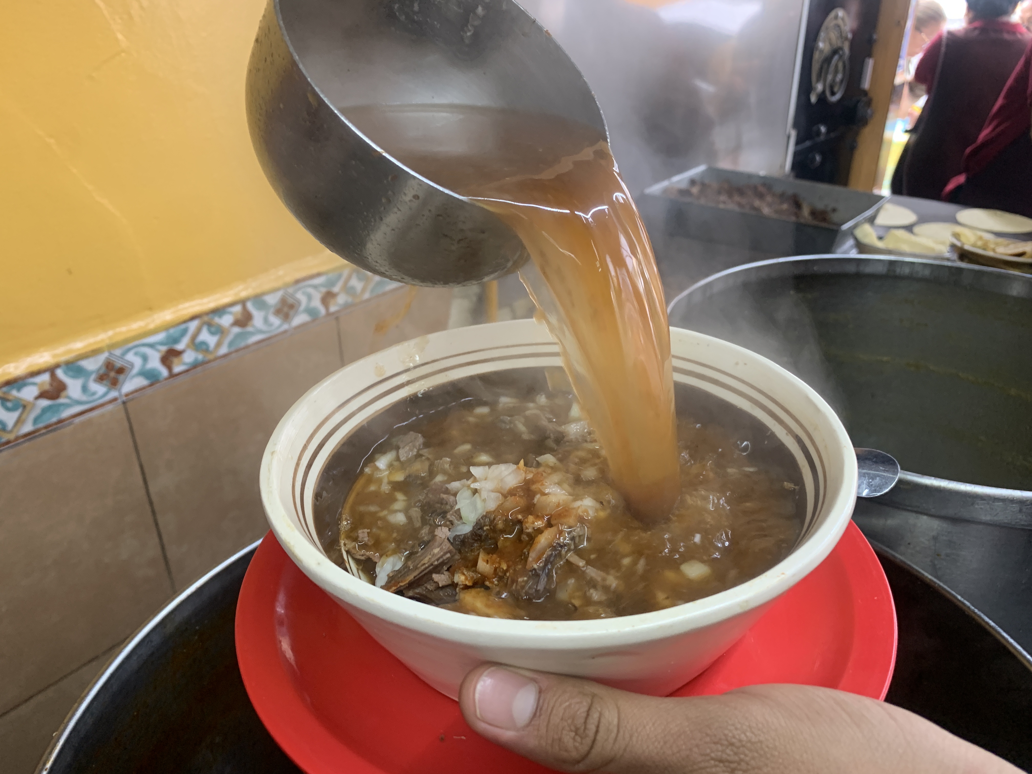 Cucharón sirviendo consomé caliente sobre un tazón de birria, con vapor, en el mostrador de la cocina