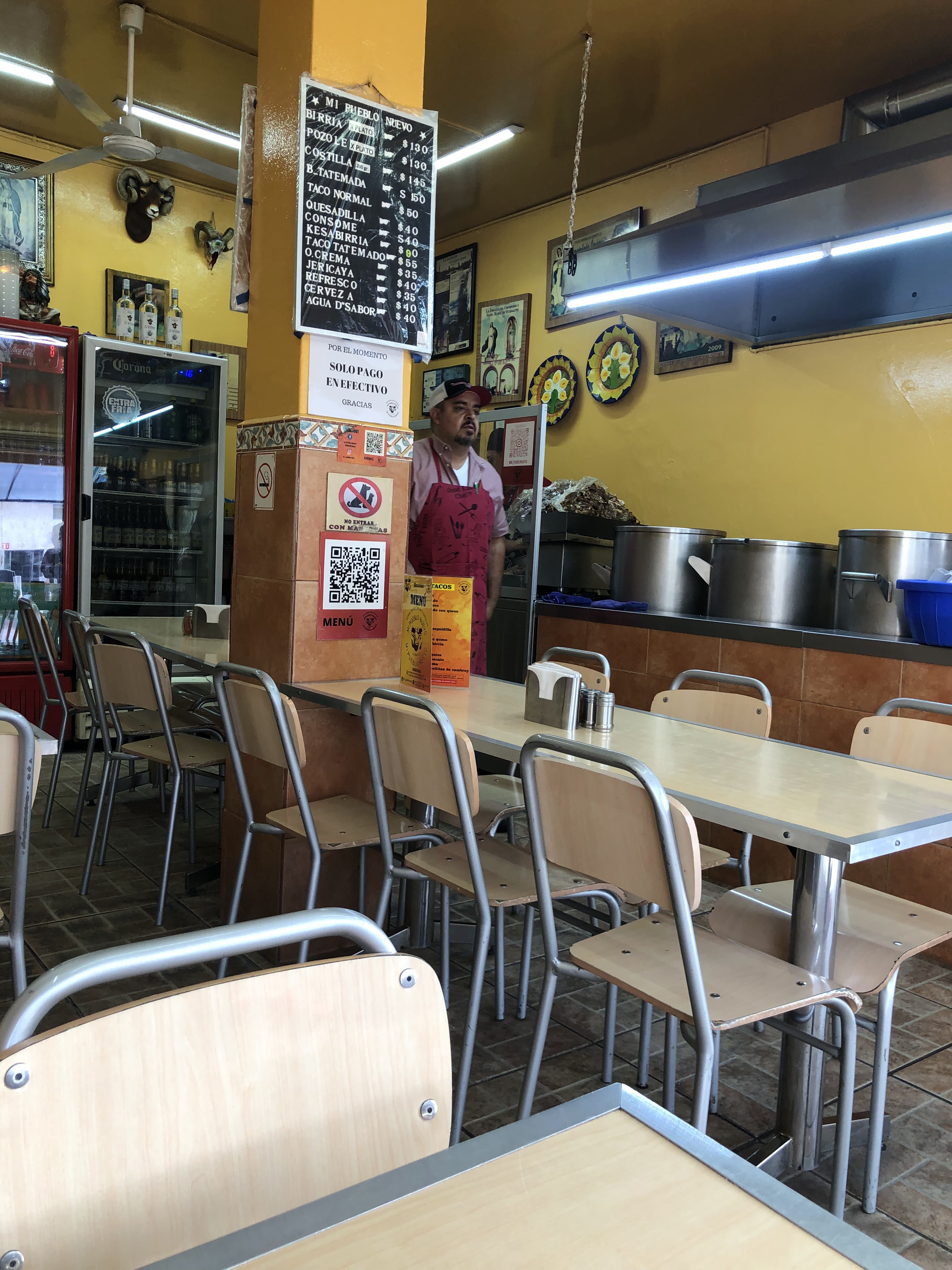 Interior del local: sillas y mesas, el pizarrón de precios en la columna y un cocinero con mandil rojo