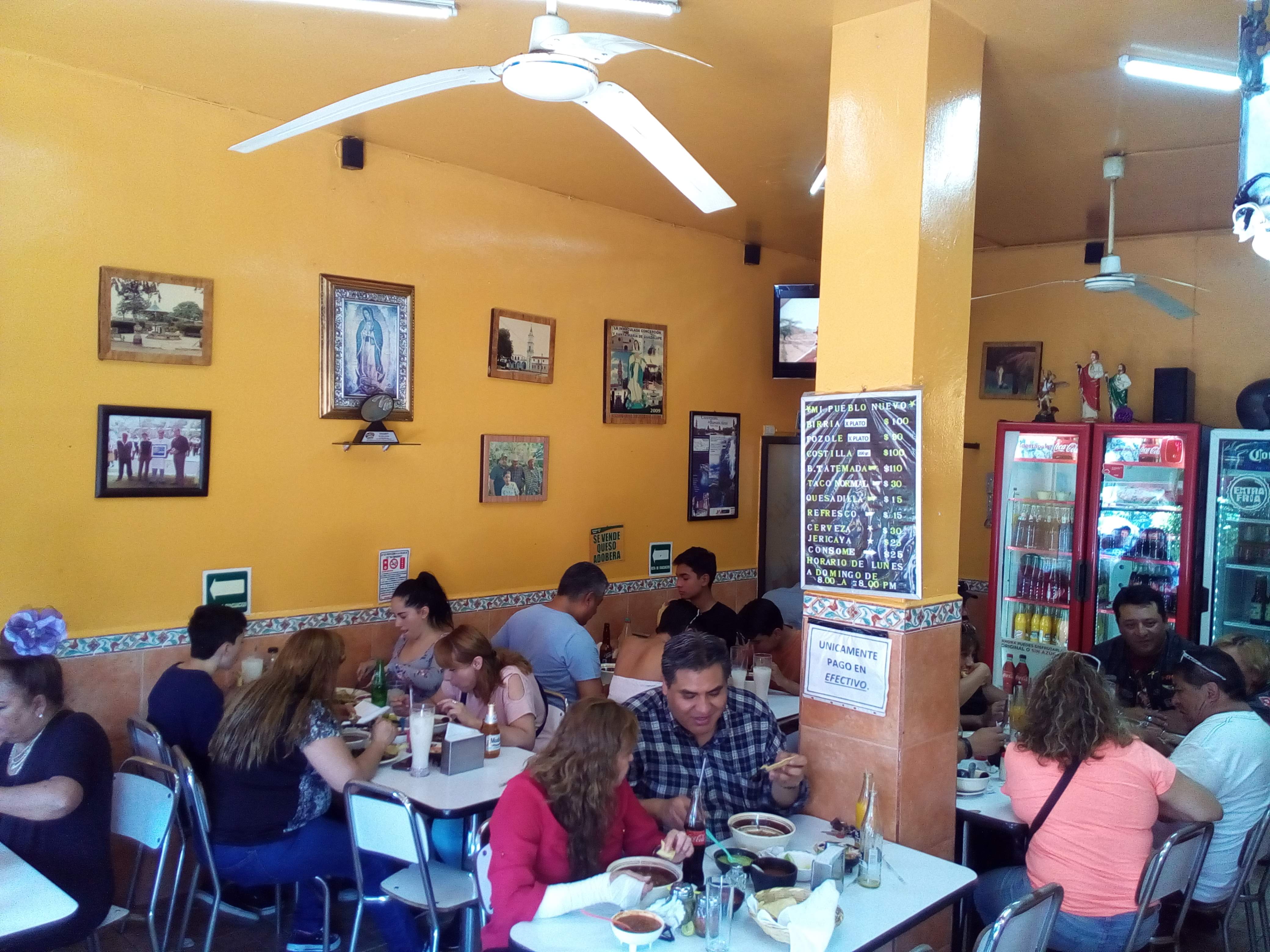 El comedor lleno a la hora de la comida, paredes amarillas, cuadros y mesas servidas con birria