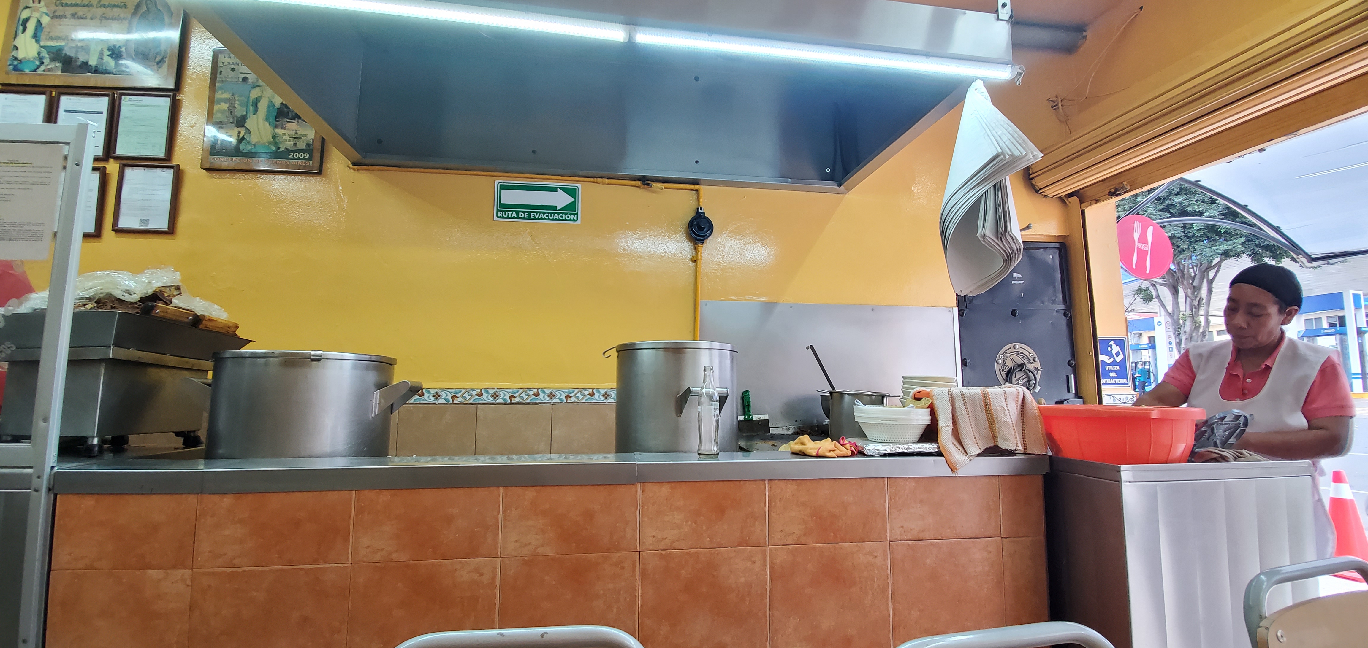La cocina vista desde las mesas: ollas de acero, campana, barra de azulejo y una cocinera trabajando