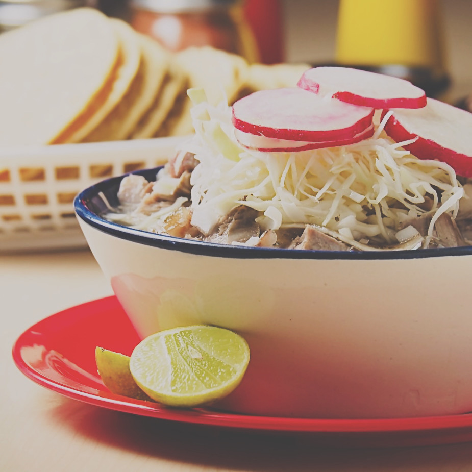 Tazón de pozole blanco con col, rábanos y limón, sobre plato rojo, con canasta de tortillas al fondo