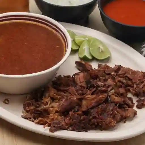 Plato de birria tatemada con queso, acompañado de consomé, limones y salsa